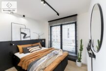 Jasna sypialnia z dużym oknem, wyposażona w łóżko z czarnym zagłówkiem i pościelą w odcieniach pomarańczu i szarości