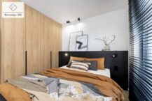 Jasna sypialnia z dużym oknem, wyposażona w łóżko z czarnym zagłówkiem i pościelą w odcieniach pomarańczu i szarości oraz w pojemną szafę w zabudowie.